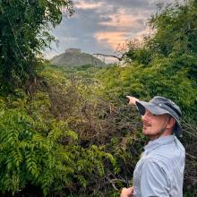 a person in a sun hat points at a mesoamerican pyramid in the background