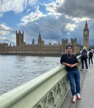 Person standing in front of the Palace of Westminster, including the iconic Big Ben, on a bridge over the River Thames.