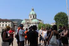 Students stand around a staff member, who is sharing about the buildings nearby in the outdoor square