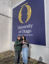 Two people standing in front of a University of Otago sign and throw their "O"