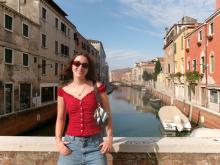 Morgan stands in a red shirt, jeans, and sunglasses, with a canal and buildings in the background