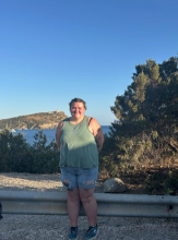 Lane stands in denim shorts and a green shirt with the ocean and rocky hills behind them