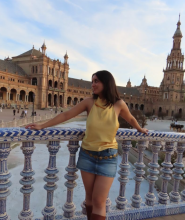 Ava stands on a balcony, with a city building in the background