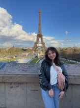 Giana smiling by the Eiffel Tower under a partly cloudy sky. 
