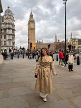 Hana smiling in front of the Big Ben in London wearing a trench coat under a cloudy sky
