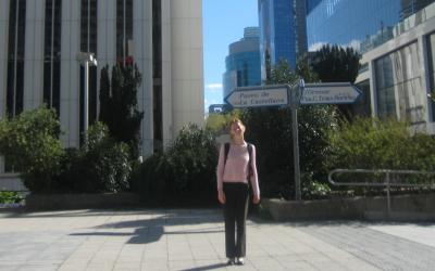 Person standing on a sunny pathway with street signs for Paseo de la Castellana and Orense in the foreground, and modern high-rise buildings in the background.