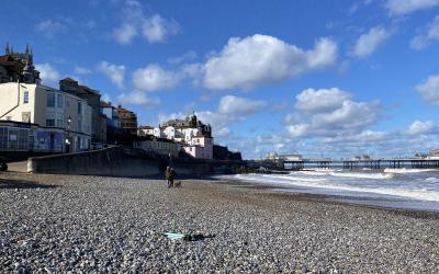 View of a pebble beach with a few individuals in the distance, facing traditional seafront buildings under a blue sky with scattered clouds.