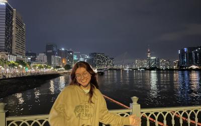 Person standing on a bridge at night with a smile, with the city skyline and a brightly lit suspension bridge in the background.