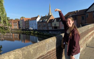 Person pretending to touch the spire of Norwich Cathedral from across the river, with colorful houses lining the opposite bank.
