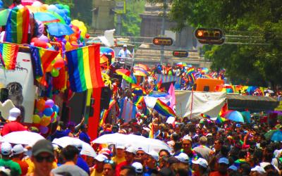 Colorful Pride parade in progress with a crowd of people and various rainbow flags and decorations in a sunny urban setting.