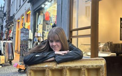A person resting their elbows on a large golden waffle statue on a street, with shops in the background.