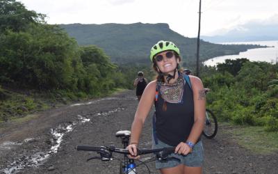 Person smiling at the camera while standing with a bicycle on a dirt path in a natural setting, wearing a helmet and sunglasses. There's a scenic view of hills and a lake in the background.