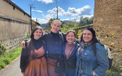 Four friends smiling and posing together on a sunny day with a scenic background of a country road and stone walls.