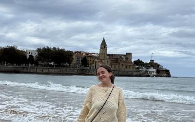 Person standing on a sandy beach with waves at their feet, smiling with the old town of Gijón, Spain, featuring the St. Peter's Church in the background under a cloudy sky.