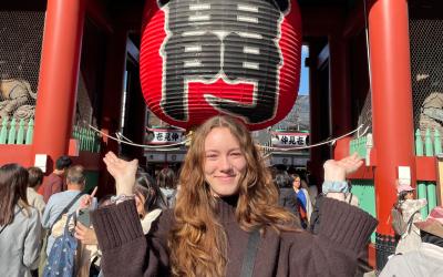 A person smiling and posing in front of the Kaminarimon gate at Senso-ji Temple, Tokyo, with a large red lantern overhead.