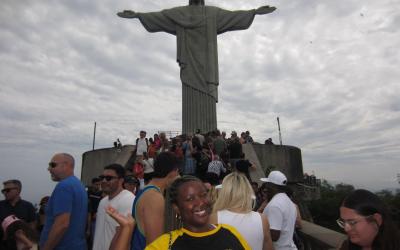 Person wearing a Brazil T-shirt smiling in front of the Christ the Redeemer statue in Rio de Janeiro, with other visitors around.
