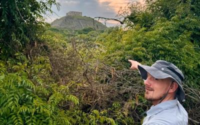 a person in a sun hat points at a mesoamerican pyramid in the background