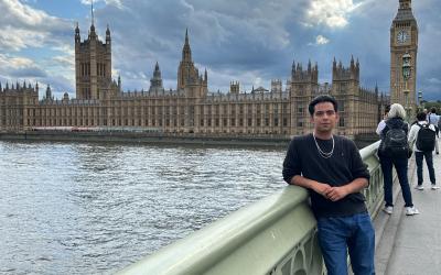 Person standing in front of the Palace of Westminster, including the iconic Big Ben, on a bridge over the River Thames.