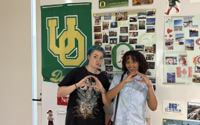 Sara and a friend standing in front of a wall of UO posters and memorabilia, throwing their "O"