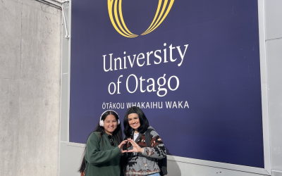 Two people standing in front of a University of Otago sign and throw their "O"