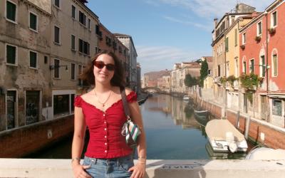 Morgan stands in a red shirt, jeans, and sunglasses, with a canal and buildings in the background