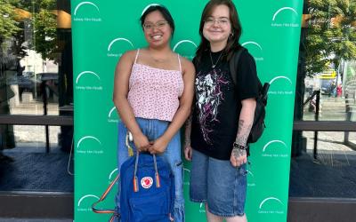 Two individuals posing in front of a promotional backdrop with a green background and white arcs