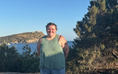 Lane stands in denim shorts and a green shirt with the ocean and rocky hills behind them