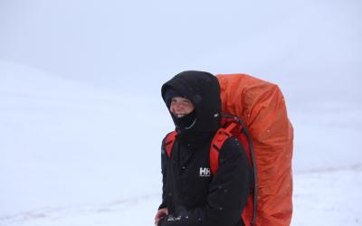 Maddy stands in a black coat with a red backpack. The background is filled with snow