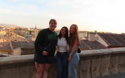 three students stand on a balcony with city buildings in the background