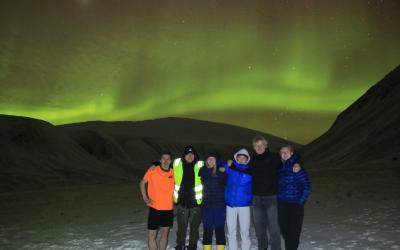 Maddy and 5 friends stand together in the snow with the northern lights tinting the sky with a green hue