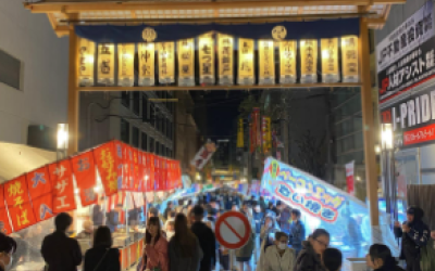 picture taken outside at nighttime; a gate above the sidewalk is illuminated by small lights, people are walking under the gate