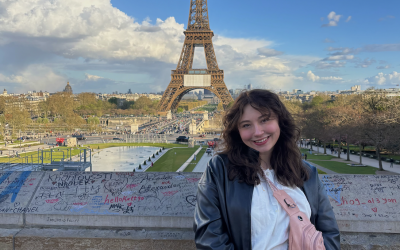 Giana smiling by the Eiffel Tower under a partly cloudy sky. 