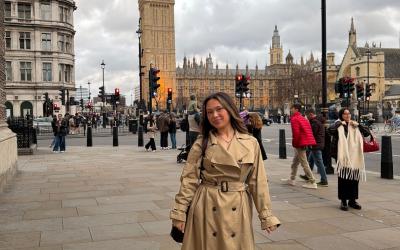 Hana smiling in front of the Big Ben in London wearing a trench coat under a cloudy sky