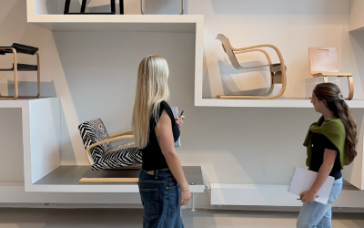 Two students walking in front of a white wall with chairs 